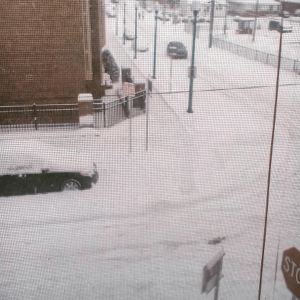 A snowy street scene, taken from a second floor apartment window. Snow covers cars, sidewalk and street. 