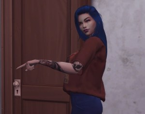 A young adult female Sim, with long blue hair and tattoos, stands in front of a white wall and wooden door, pointing to something out o frame. 