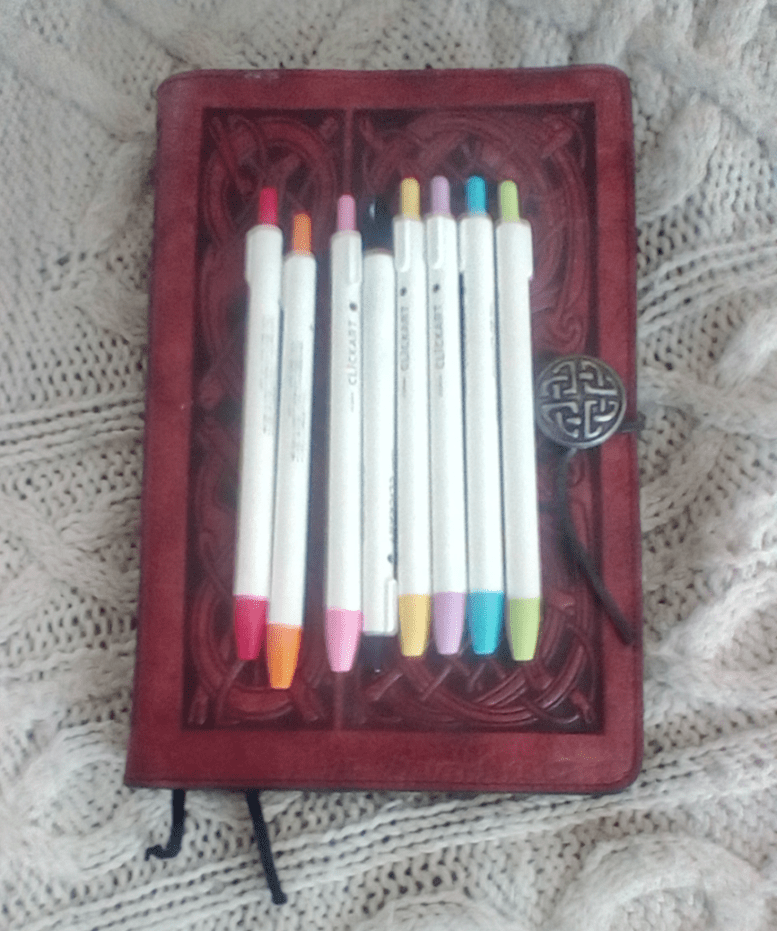 Eight Clickart markers, in summery bright colors, rest on top of a Celtic-tooled leather journal cover. 