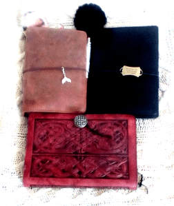 Three leather/leather look notebook covers, in shades of brown and black. The black one had a metal tag reading "Not all who wander are lost" by C.S. Lewis. The light brown on has a mermaid tail charm, and the largest has tooled leather in a design that coordinates with the celtic knotwork metal button. 